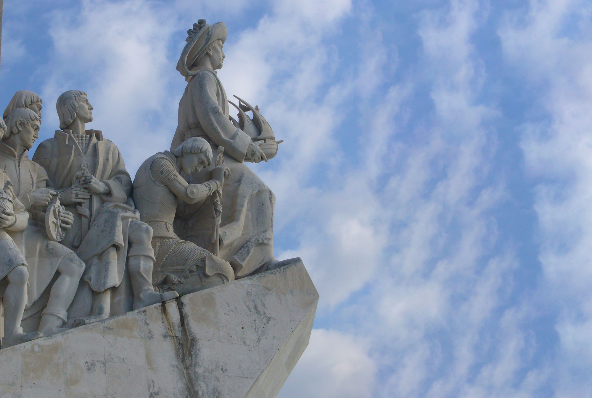 Monument to Magellan in Lisbon, Portugal. The explorer stands on a promontory, looking out into a blue sky filled with clouds.